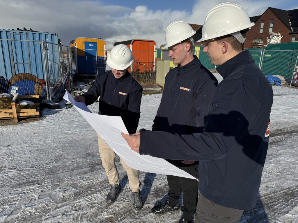 Drie vakmensen van Bouwsector Nederland met witte helm bekijken een bouwtekening op een besneeuwde bouwplaats